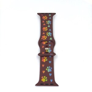May include: A brown silicone watch band featuring a pattern of colorful paw prints and hearts. The paw prints are in shades of orange, yellow, green, and blue. The band has a buckle closure and is designed for a smartwatch.
