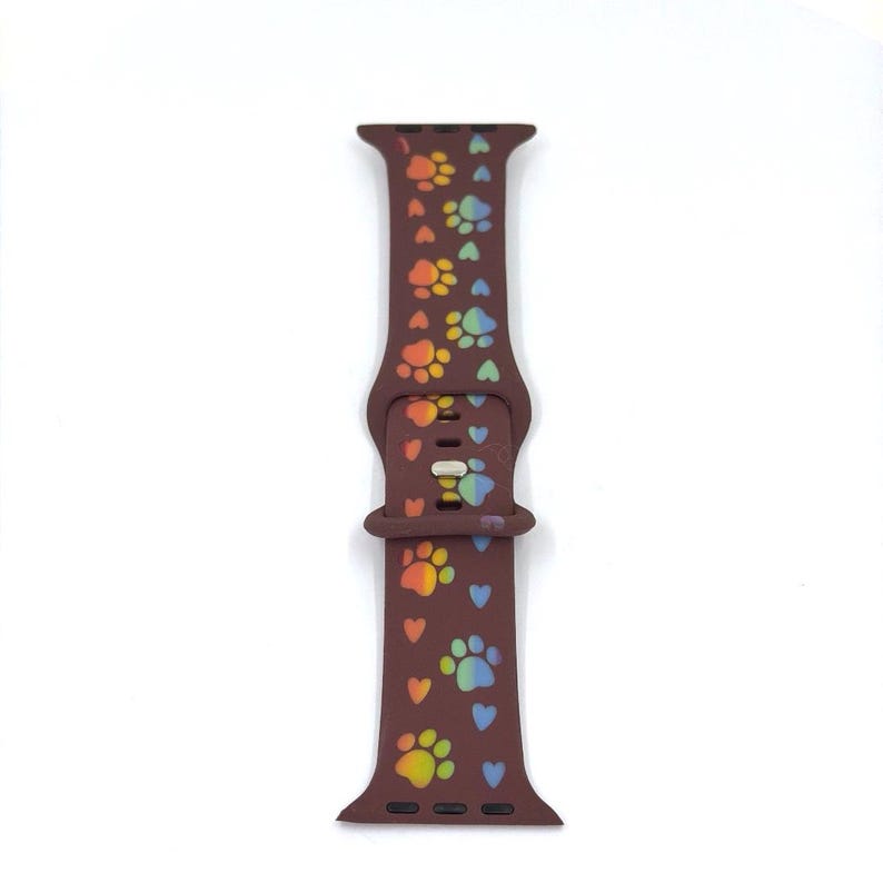 May include: A brown silicone watch band featuring a pattern of colorful paw prints and hearts. The paw prints are in shades of orange, yellow, green, and blue. The band has a buckle closure and is designed for a smartwatch.