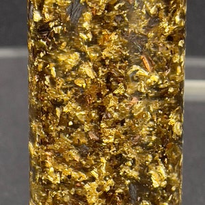 May include: A close-up of a cylindrical sample of a dark green material with flecks of gold throughout.