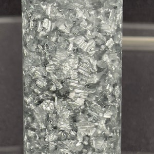 May include: A clear glass cylinder filled with silver glitter. The glitter is densely packed and creates a shimmering effect.