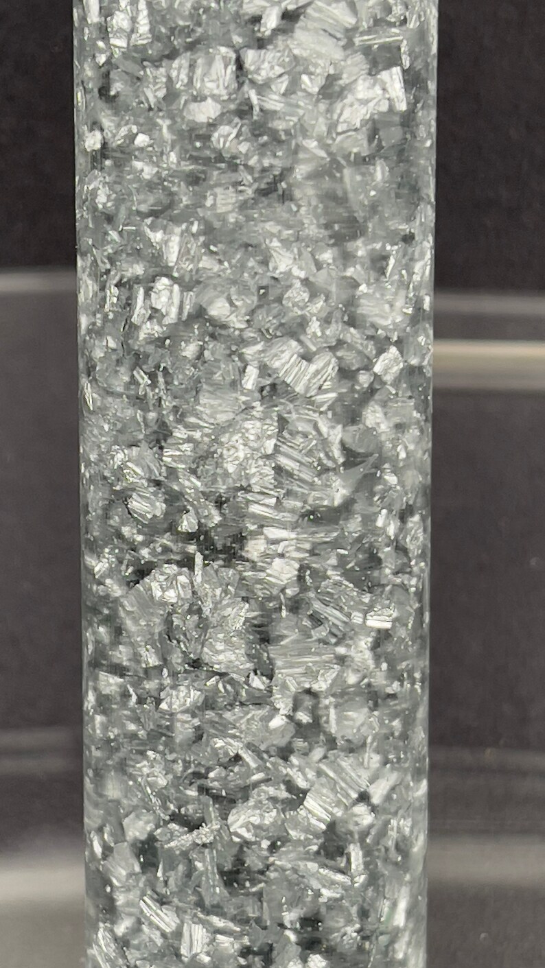 May include: A clear glass cylinder filled with silver glitter. The glitter is densely packed and creates a shimmering effect.