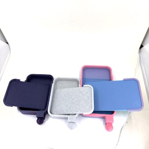 May include: Three small, rectangular, silicone cases with lids. The cases are in different colors: navy blue, light gray, and pink. The lids are open and the cases are lined with a soft, gray material.