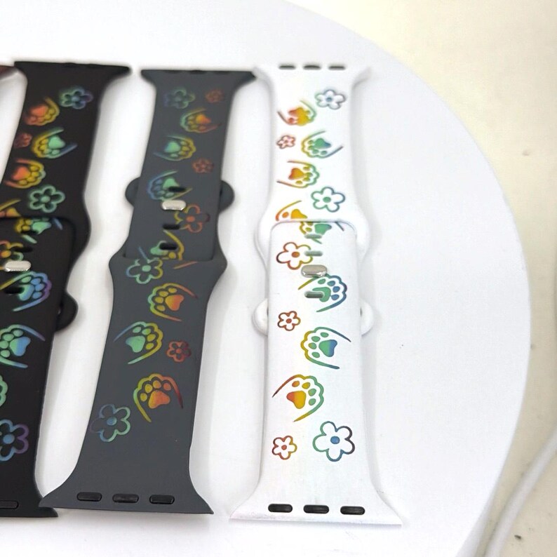 May include: Three watch bands in black, gray, and white. Each band features a repeating pattern of paw prints and flowers in rainbow colors. The bands are made of a flexible material and have a secure clasp.