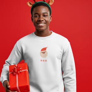 Puede incluir: Una sudadera blanca con un gráfico de Papá Noel y el texto "Ho Ho Ho". La persona lleva una diadema de cuernos de reno y sostiene una caja de regalo roja con un lazo.