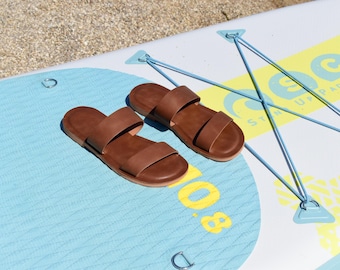 May include: A pair of brown leather sandals with two straps, resting on a light blue and white paddleboard. The paddleboard has yellow and blue accents and the number "8.0" in yellow. The sandals are placed on the board near the beach.