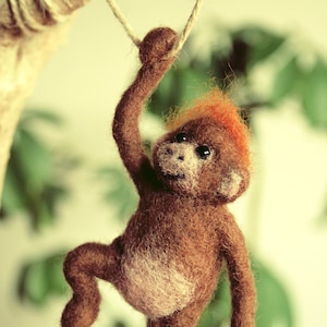 Può includere: Un giocattolo di scimmia di feltro marrone e arancione appeso a una corda. La scimmia tiene la corda con una mano e l'altra mano pende. La scimmia ha una macchia bianca sul petto.