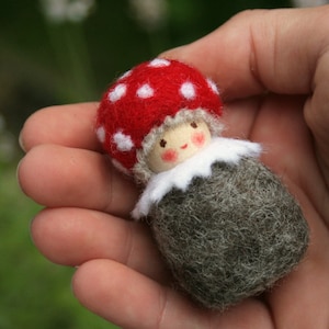 Könnte beinhalten: Ein kleiner Filz-Gnome mit einem rot-weiß gepunkteten Hut und einem grauen Filzkörper. Der Gnome hat ein bemaltes Gesicht mit rosigen Wangen.