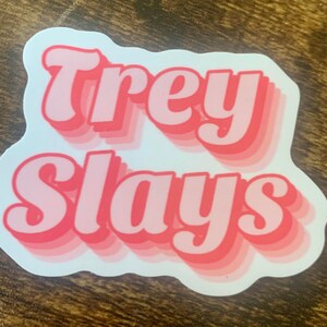 May include: Pink and white sticker with the text "Trey Slays" in a retro font.