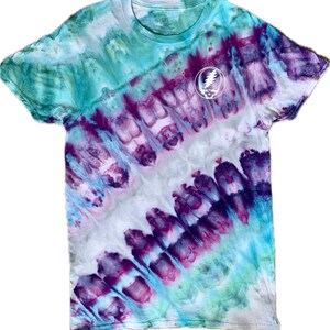 Puede incluir: Una camiseta tie-dye con un patrón de remolino morado, azul y verde. La camiseta tiene un círculo blanco con un diseño de calavera y rayo en el centro.