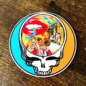 May include: A round sticker featuring a colorful psychedelic image of a skull with a mouth open wide, surrounded by a blue and orange background. The skull is a popular symbol of the Grateful Dead band.