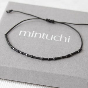 Puede incluir: Una pulsera minimalista de cuentas negras con cierre de nudo corredizo, colocada sobre una caja gris. La caja tiene la palabra "mintuchi" impresa. La pulsera presenta pequeñas cuentas oscuras y detalles plateados.
