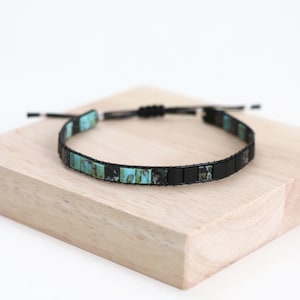 Peut inclure: Un bracelet en perles noires et turquoise avec une fermeture à nœud coulissant. Le bracelet est composé de petites perles carrées et est posé sur une surface en bois.