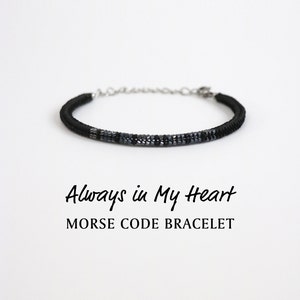 May include: A black and grey beaded bracelet with a silver clasp. The bracelet has the words "Always in My Heart" written below it, and the words "Morse Code Bracelet" written below that.