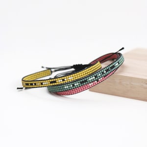 Puede incluir: Dos pulseras de cuentas con patrones geométricos coloridos. La pulsera superior es amarilla con detalles en blanco y negro. La pulsera inferior es rosa, verde y blanca.