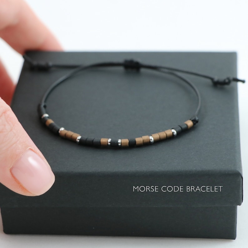 Peut inclure: Un bracelet en cordon noir avec des perles carr&eacute;es noires, argent&eacute;es et brunes. Le bracelet est dans une bo&icirc;te noire avec les mots "Bracelet Code Morse" imprim&eacute;s sur le devant.