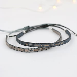 Könnte beinhalten: Zwei Perlenarmbänder, eines dunkelblau und eines hellgrau mit goldenen Akzenten. Die Armbänder sind mit einer dunkelblauen Schnur zusammengebunden.