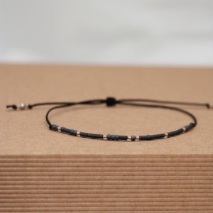 Op de afbeelding: Een dunne zwarte koordarmband met een reeks kleine, matte donkergrijze en metallic goudkleurige cilindrische kralen in een patroon. De armband is voorzien van een verstelbare schuifknoopsluiting en twee losse uiteinden met kleine zilverkleurige kralen.