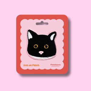 May include: An embroidered iron-on patch featuring a black cat face with white inner ears, yellow eyes, and a pink nose. The patch is mounted on a scalloped-edge pink card with red borders, labelled Iron on Patch and TheUrbanite.