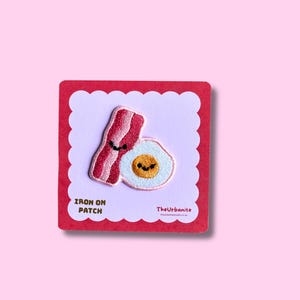 Puede incluir: Un parche bordado termoadhesivo que muestra una tira de tocino y un huevo frito, ambos con pequeños ojos negros y sonrisas. El parche está sujeto a una tarjeta cuadrada con un borde blanco festoneado y bordes rojos, etiquetada IRON ON PATCH y TheUrbanite.