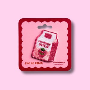 Op de afbeelding: Een geborduurde strijkpatch in de vorm van een aardbeienmelkpak in roze en witte tinten. De patch bevat een kleine groene aardbeigrafiek en de tekst 'STRAWBERRY MILK' op een donkerrode achtergrondkaart met geschulpte randen.