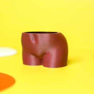 K&ouml;nnte beinhalten: Ein brauner Keramik-Pflanztopf in Form eines menschlichen Torsos. Der Topf steht auf einer gelben Oberfl&auml;che.