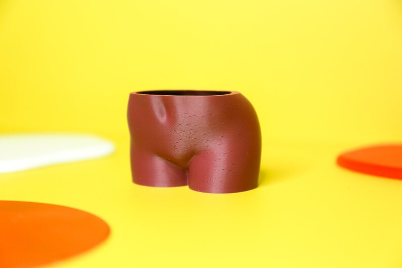 K&ouml;nnte beinhalten: Ein brauner Keramik-Pflanztopf in Form eines menschlichen Torsos. Der Topf steht auf einer gelben Oberfl&auml;che.