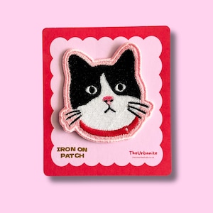 May include: Embroidered patch featuring a black and white cat face with a pink nose and red collar. The patch is on a pink scalloped background, atop a red rectangular card. Text on the card reads "IRON ON PATCH" and "TheUrbanite".