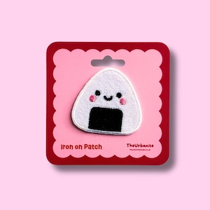 Peut inclure: Un écusson thermocollant en forme de triangle représentant un onigiri blanc avec une base noire, deux yeux noirs, un petit sourire et des joues roses. L'écusson est fixé sur un carton rouge et rose avec le texte Iron on Patch et TheUrbanite.