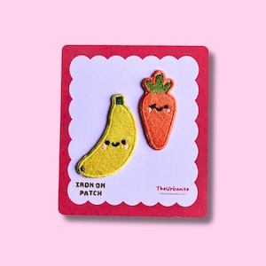 Peut inclure: Deux écussons thermocollants brodés représentant une banane jaune et une carotte orange, toutes deux avec des visages souriants et des joues roses. Les écussons sont fixés sur une carte blanche festonnée à bord rouge, marquée IRON ON PATCH et TheUrbanite.