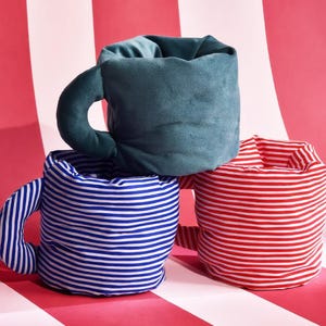 May include: Three plush mug-shaped cushions. One is solid dark teal velvet, one features blue and white horizontal stripes, and one features red and white horizontal stripes. Each soft, stuffed item includes a curved handle on the side.