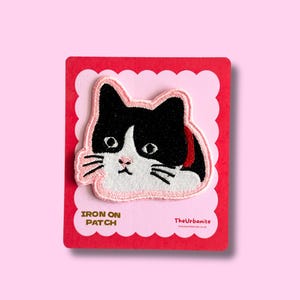 May include: Embroidered iron-on patch of a black and white cat face with pink detailing. The patch is on a red card with the text "IRON ON PATCH" and the brand name "TheUrbanite". The background is a pale pink.