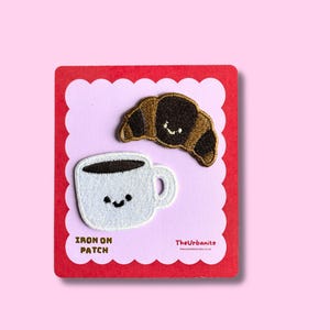 May include: Two embroidered iron-on patches featuring a brown croissant and a white coffee mug, both with simple black facial features. The patches are displayed on a scalloped-edge card with the text IRON ON PATCH and TheUrbanite.