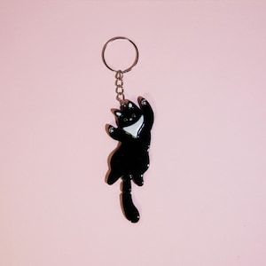 May include: Black cat keychain with a white chest and paws, hanging from a silver chain and ring. The cat is in a playful pose, with its arms outstretched. The background is a solid light pink.
