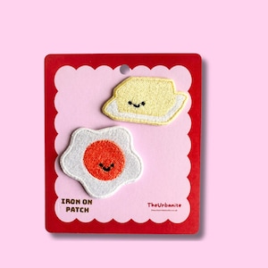 Puede incluir: Dos parches bordados termoadhesivos con un huevo frito sonriente y una porción de mantequilla sonriente. Los parches se presentan sobre una tarjeta de soporte festoneada roja y rosa claro con el texto IRON ON PATCH y TheUrbanite impreso.