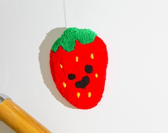 May include: A red and green felt strawberry with a smiling face hanging from a white string. The strawberry has yellow seeds and a black outline.