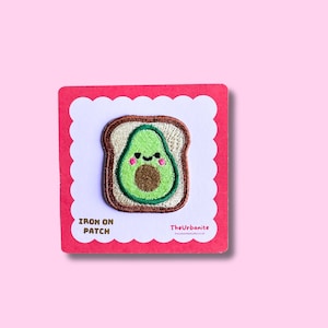 May include: An embroidered iron-on patch featuring a cartoon avocado on a slice of toast with a smiling face, pink cheeks, and a brown pit. The patch is mounted on a square red card with white scalloped edges, labeled IRON ON PATCH and TheUrbanite.