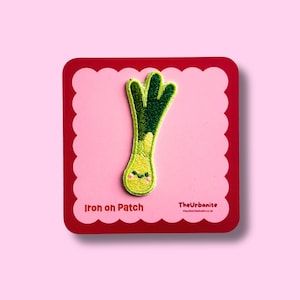 May include: An embroidered iron-on patch shaped like a leek with a smiling face, featuring shades of light and dark green. The patch is mounted on a square pink card with scalloped edges, displaying the text Iron on Patch and TheUrbanite.