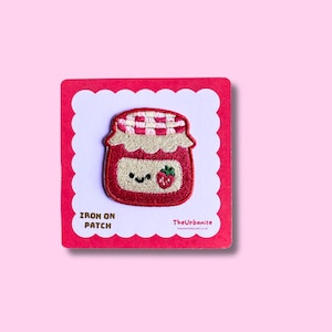 May include: An embroidered iron-on patch featuring a red jam jar with a white and red checkered lid. The jar has a cream-colored label with a smiling face and a small red strawberry. The patch is mounted on a square card labeled IRON ON PATCH.