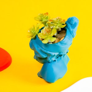 May include: A blue ceramic planter shaped like a bust, with a succulent plant growing inside. The planter is on a yellow surface.