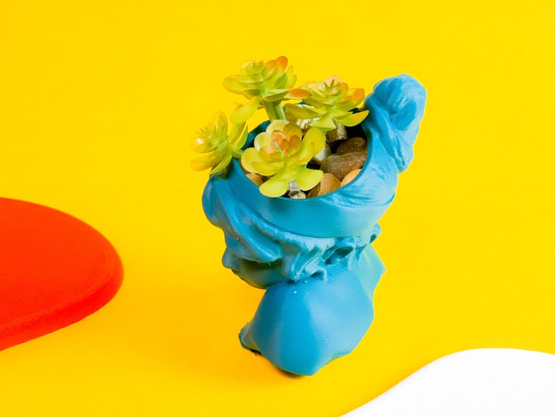 May include: A blue ceramic planter shaped like a bust, with a succulent plant growing inside. The planter is on a yellow surface.