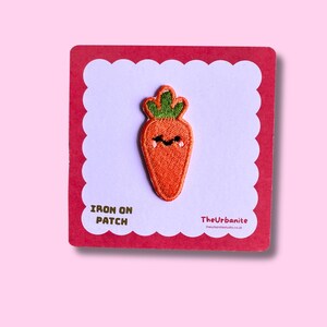 May include: An embroidered iron-on patch shaped like an orange carrot with a green leafy top and a smiling face with pink cheeks. The patch is mounted on a square white card with a scalloped edge and red border, featuring the text IRON ON PATCH.