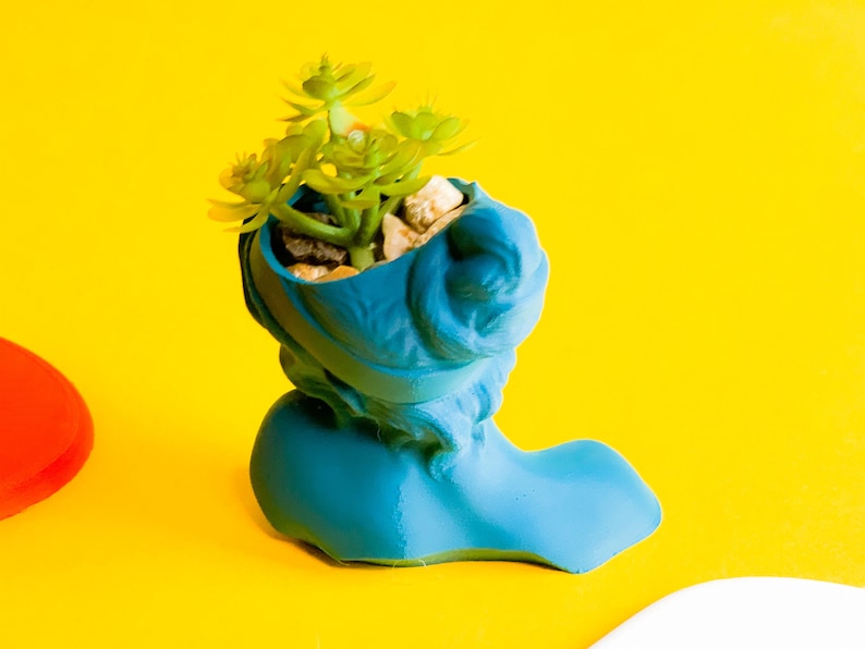 May include: A blue ceramic planter shaped like a shoe with a small succulent plant growing inside. The planter is sitting on a yellow surface.