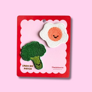 May include: Two embroidered patches on a red scalloped card. One patch is a green broccoli floret with a smiling face. The other is a fried egg with a smiling face. The text "IRON ON PATCH" is below the broccoli.