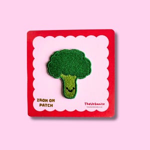 May include: A green broccoli iron-on patch with a smiling face, on a red and white scalloped-edge card. The text "IRON ON PATCH" is visible. TheUrbanite logo is also present. The background is a solid light pink.