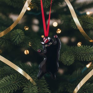 May include: A black cat Christmas tree decoration with a red ribbon, hanging on a Christmas tree. The cat is wearing a party hat and has red paws. Gold ribbon and Christmas lights are in the background.