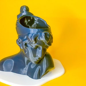 May include: A gray bust sculpture of a person with a broken top, resembling a planter. The bust is sitting on a white surface against a yellow background.
