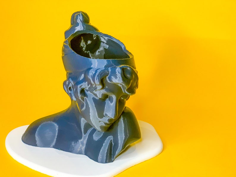 May include: A gray bust sculpture of a person with a broken top, resembling a planter. The bust is sitting on a white surface against a yellow background.