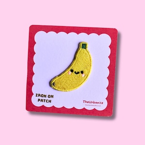 Può includere: Una toppa termoadesiva a forma di banana gialla con un viso sorridente, guance rosa e un gambo verde. La toppa è fissata su un cartoncino quadrato con bordo rosso e centro bianco smerlato, con il testo IRON ON PATCH e TheUrbanite.