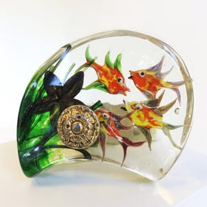 May include: A clear, curved glass paperweight featuring colorful fish and aquatic plants. The fish are orange, yellow, and purple, with green accents. A decorative, gold-toned button is embedded in the glass, adding a vintage touch.