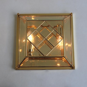 May include: A square, gold-toned wall sconce with a geometric design. The sconce features a mirrored interior and two exposed light bulbs. The fixture is mounted on a white wall, creating a contrast with the warm gold tones.